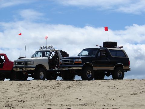 1996 Ford Bronco - Several off-road vehicles are parked on a sandy dune under a partly cloudy sky, with red flags flying.