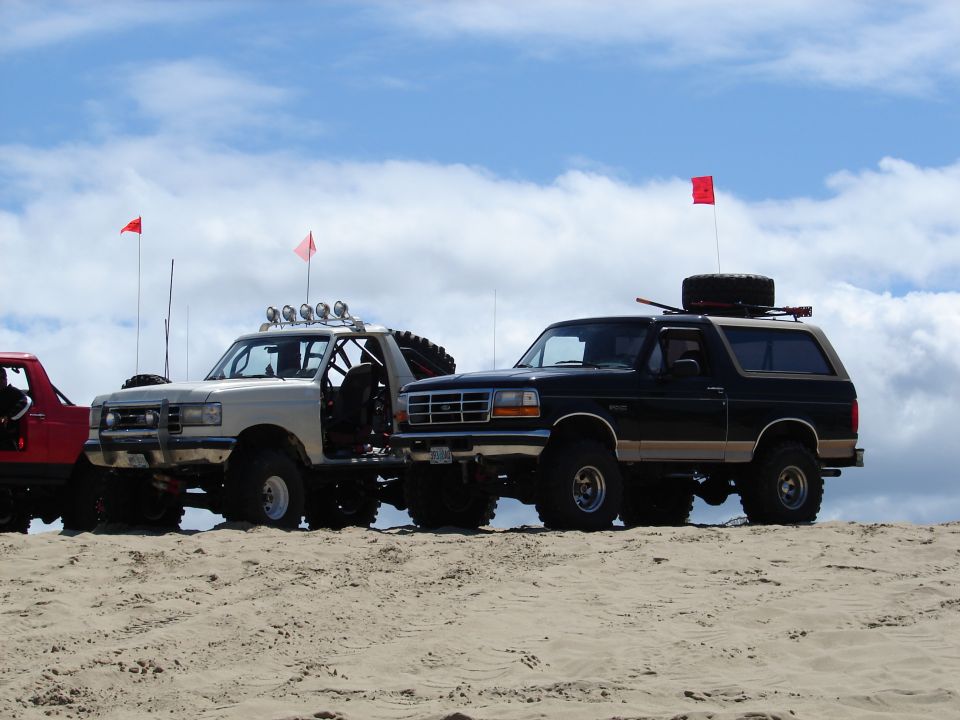 Two modified SUVs parked on sand dunes under a cloudy sky. One is white with a roll cage, the other is black with a spare tire on top