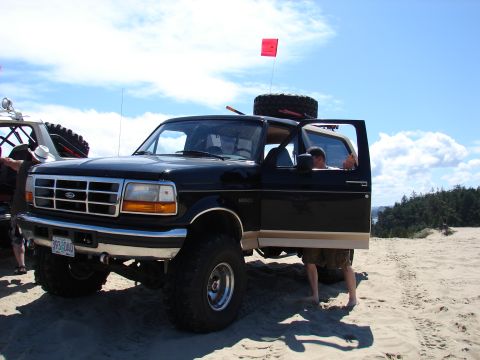 1996 Ford Bronco - A black 1996 Ford Bronco with tan trim sits on a sandy beach with the driver's door open and a spare tire mounted on the roof.