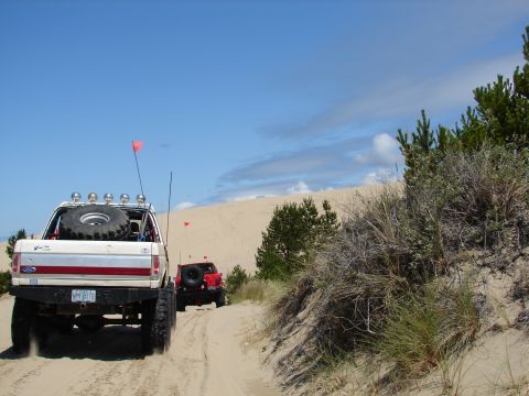 1996 Ford Bronco - A white Ford Bronco with a red stripe and a spare tire on the back drives on sand dunes, with another vehicle following behind.