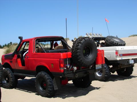 1996 Ford Bronco - A red 1996 Ford Bronco and a white Ford truck are parked on a sandy dune under a clear blue sky.