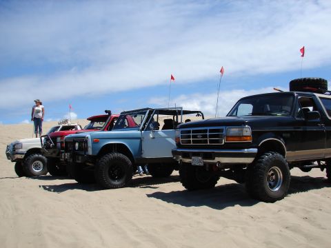 1996 Ford Bronco - Four modified SUVs, including a 1996 Ford Bronco, are parked on sand dunes on a sunny day.