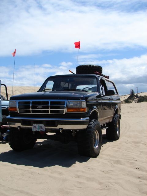 1996 Ford Bronco - A lifted black Ford Bronco is parked on a sandy dune under a cloudy sky, with red flags visible.