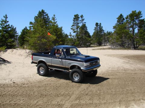 1996 Ford Bronco - A lifted two-tone Ford Bronco navigates sandy terrain under a clear blue sky.