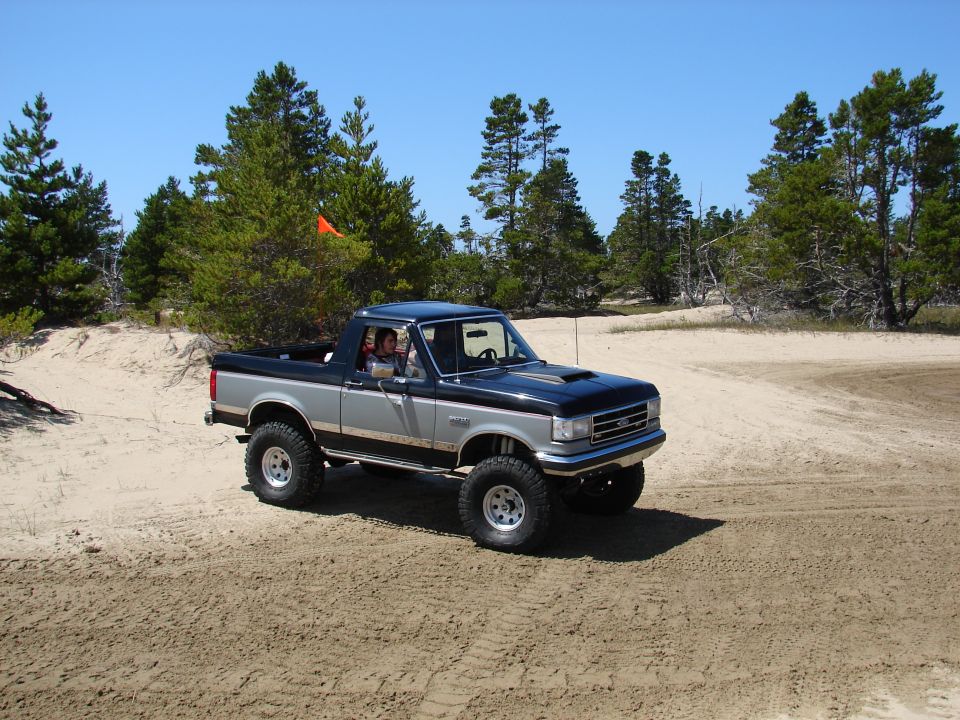 Two-tone black and silver lifted Ford Bronco with aftermarket hood scoop driving on sand dunes