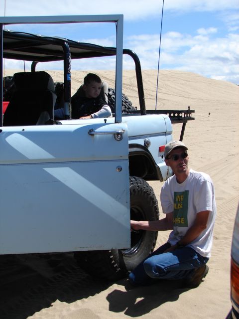 1996 Ford Bronco - A man checks the tire of a light blue 1978 Ford Bronco while a child sits in the passenger seat, with sand dunes in the background.