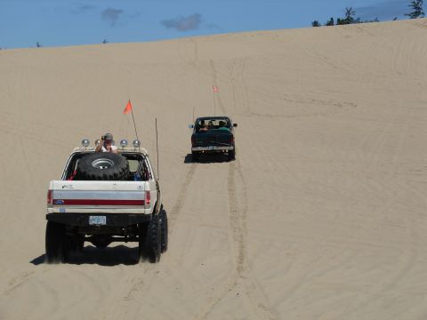 1996 Ford Bronco - Two trucks drive through sand dunes under a blue sky.