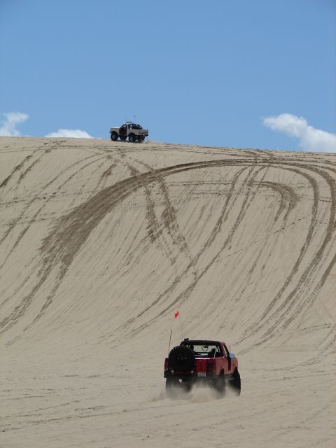 1996 Ford Bronco - A red 1996 Ford Bronco navigates sandy terrain, with another vehicle visible on the crest of a dune.