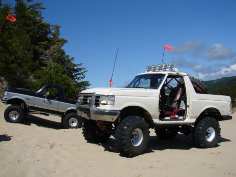 1996 Ford Bronco - Two modified trucks are parked on a sandy dune area under a clear blue sky.