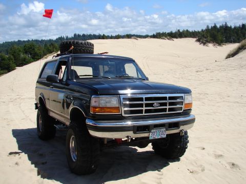 1996 Ford Bronco - A black 1996 Ford Bronco is parked on a sandy terrain with a spare tire mounted on its roof and a red flag flying.