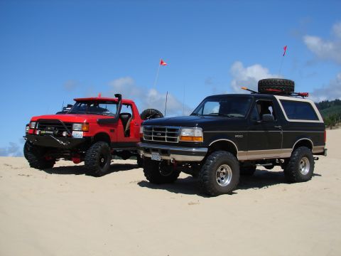 1996 Ford Bronco - Two off-road vehicles, a black Ford Bronco and a red truck, sit on sand dunes on a sunny day.
