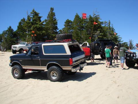 1996 Ford Bronco - Several off-road vehicles are gathered on a sandy beach with trees in the background under a clear blue sky.