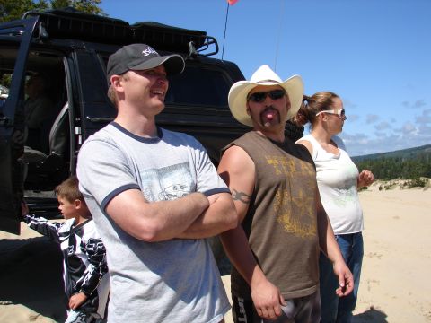 1996 Ford Bronco - People stand near a black SUV with a roof rack on a sandy terrain under a clear blue sky.