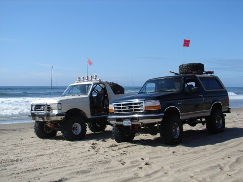 1996 Ford Bronco - Two Ford Broncos are parked on the sand near the ocean, both equipped with off-road modifications and flags.