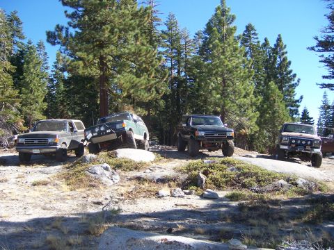 1991 Ford Bronco - Four off-road SUVs are parked on a rocky, wooded hillside under a clear blue sky.