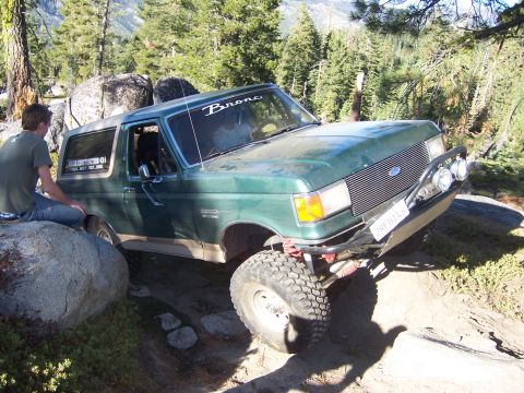 1991 Ford Bronco - A green Ford Bronco navigates a challenging off-road trail with large rocks and trees surrounding it.
