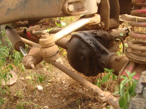 1992 Ford Bronco - Low-angle underside view of a dust-covered solid front axle showing the cast differential housing, drag link and tie rod with tapered ends,…
