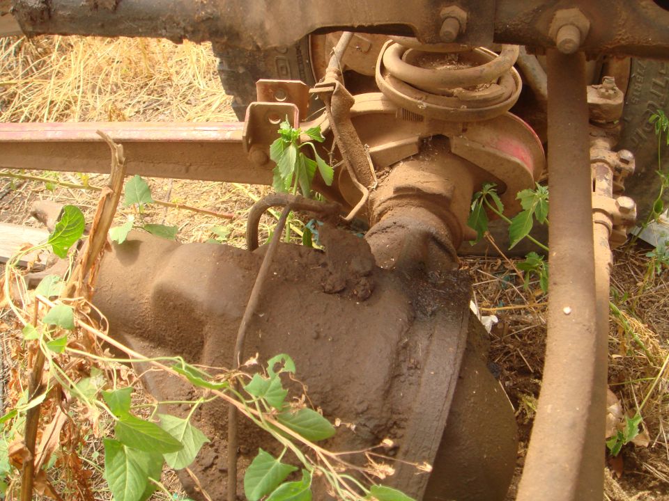 Close-up of a muddy vehicle differential and suspension components with weeds growing around them