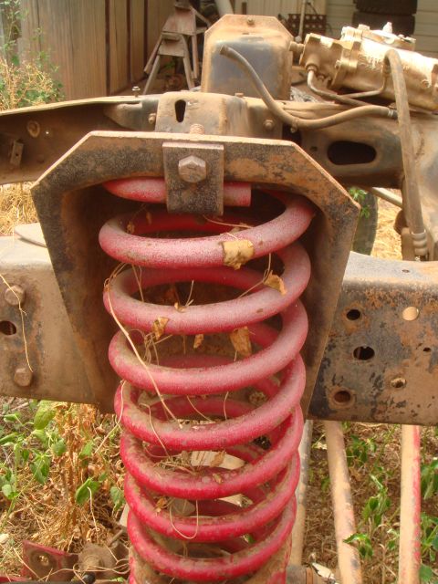 1992 Ford Bronco - A close-up view of a red suspension coil spring on a rusty vehicle frame, with dry leaves and debris caught in the coils.