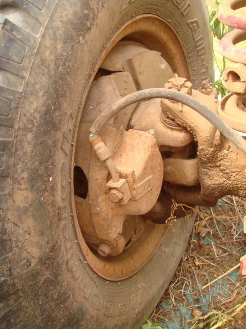 1992 Ford Bronco - Close-up of a rusty, mud-coated front axle knuckle, disc brake caliper and rotor still inside a BFGoodrich tire on a steel wheel, photograp…
