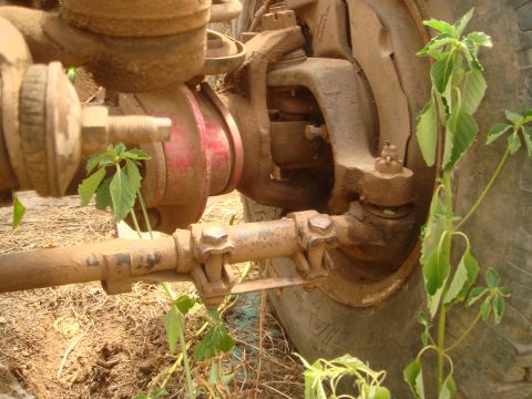 1992 Ford Bronco - Mud-covered front axle steering components and tire, with weeds growing around.