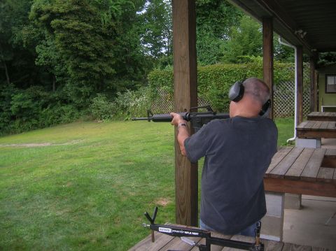 1996 Ford Bronco - Man wearing ear protection aims a rifle at an outdoor shooting range with grassy field and trees in the background.