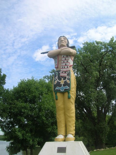 1996 Ford Bronco - A large roadside statue depicts a Native American man holding a rifle, dressed in traditional attire with yellow pants and a beaded vest.