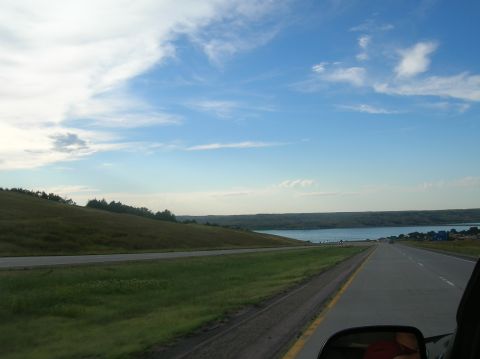 1996 Ford Bronco - A scenic drive unfolds along a highway, with rolling hills and a vast lake stretching towards the horizon under a bright, cloudy sky.