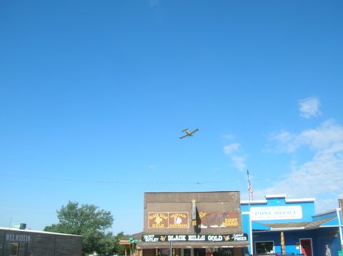 1996 Ford Bronco - A small yellow airplane flies in the sky above a street lined with shops and a post office.