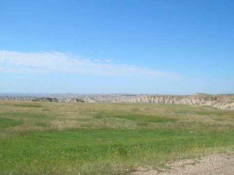 1996 Ford Bronco - A wide expanse of green grass stretches towards a dramatic badlands landscape under a bright blue sky.
