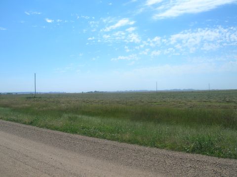 1996 Ford Bronco - A wide expanse of grassy field stretches to the horizon under a bright blue sky with wispy clouds, bordered by a dirt road.