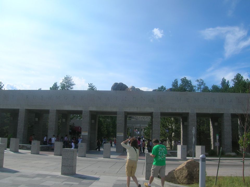 Stone archway with "MOUNT RUSHMORE NATIONAL MEMORIAL" carved above, people walking through arches under a blue sky