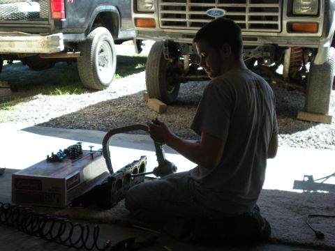1985 Ford Bronco - Mechanic works on engine components with a Ford truck visible in the background.