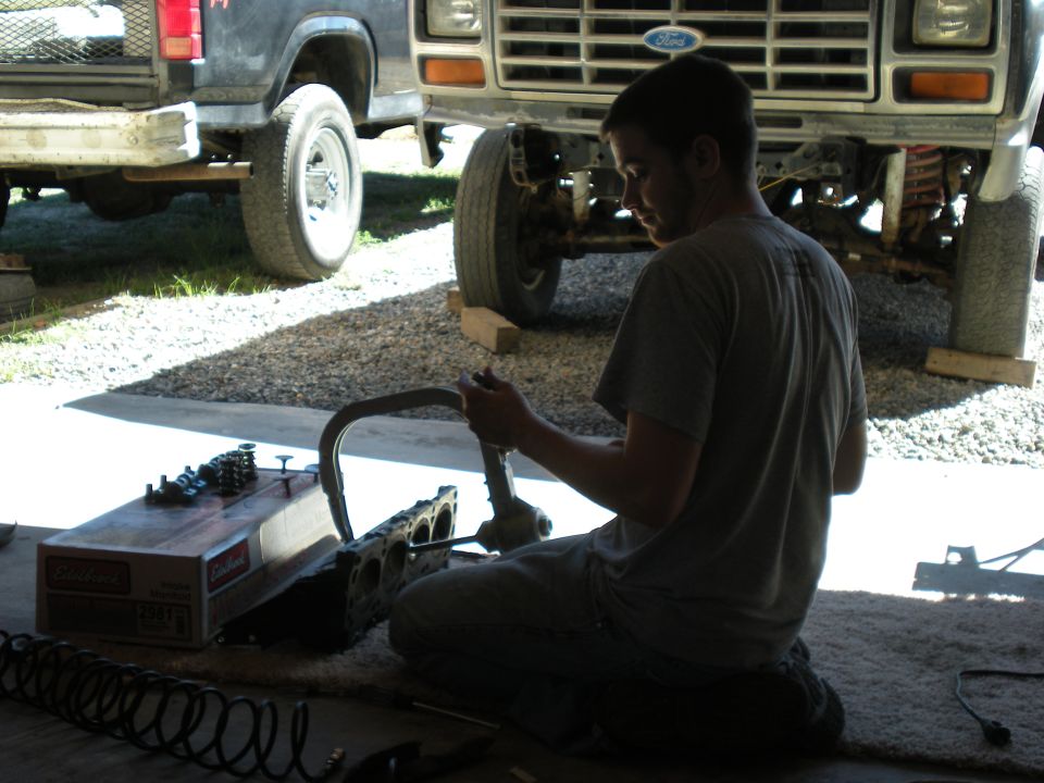 man kneeling on ground working on engine block and valve train parts, with a Ford truck in the background