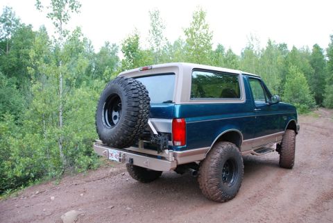 1996 Ford Bronco - Rear view of a 1996 Ford Bronco, equipped with a spare tire, on a dirt road surrounded by trees.