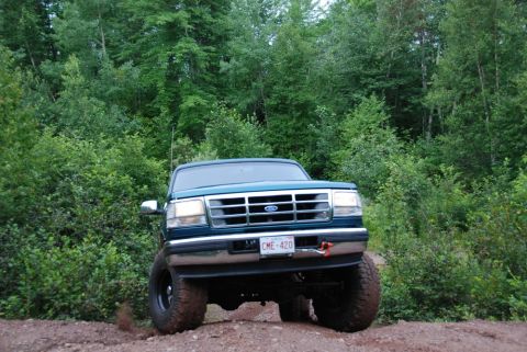 1996 Ford Bronco - A green 1996 Ford Bronco is shown from the front, driving on a dirt path with trees in the background.