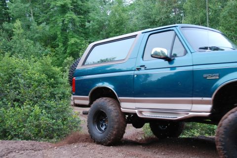 1996 Ford Bronco - This teal Ford Bronco is driving on a dirt road surrounded by trees. The spare tire is mounted on the rear door.