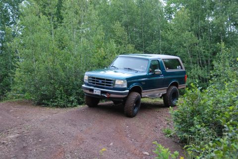 1996 Ford Bronco - A teal 1996 Ford Bronco with a tan top drives on a dirt road surrounded by trees.