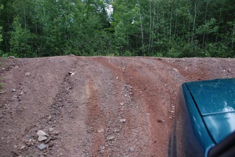 1996 Ford Bronco - A teal vehicle navigates a rough dirt road with visible tire tracks, surrounded by trees.