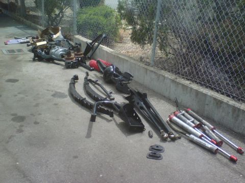 1984 Ford Bronco - Various vehicle suspension and drivetrain parts are spread across a concrete surface next to a chain-link fence.
