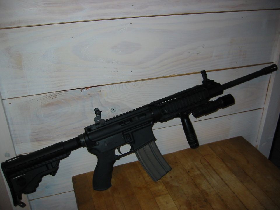 Black assault rifle with a magazine, vertical foregrip, and tactical light, resting on a wooden surface against a white wooden wall