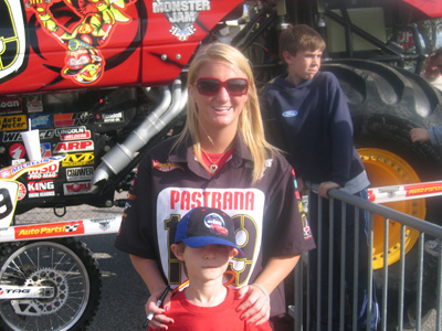 1987 Ford Bronco - A monster truck with "Monster Jam" branding is visible behind a group of people, including a woman wearing a "Pastrana" shirt.