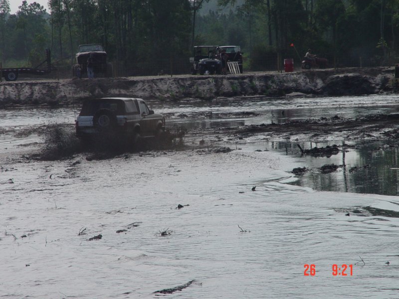 Full-size SUV with rear spare crossing a flooded mud bog as spectators watch from the bank