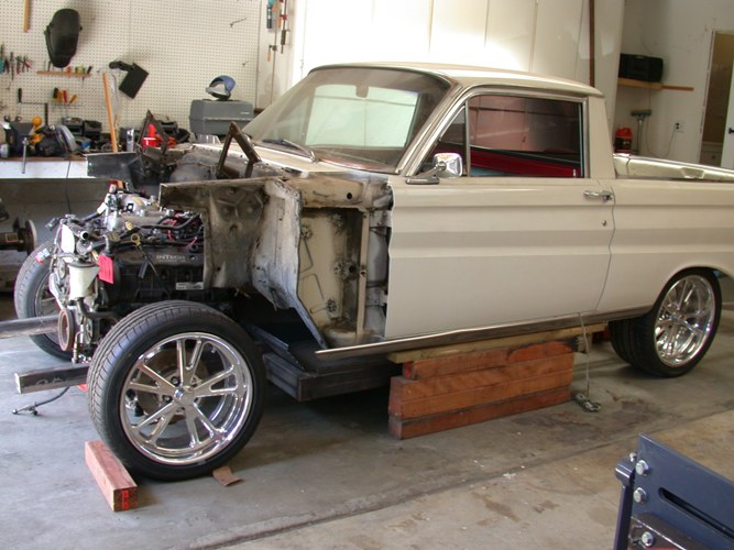 Tan 1960s Ford Ranchero mid-restoration on blocks in garage, front fender off, chrome wheels