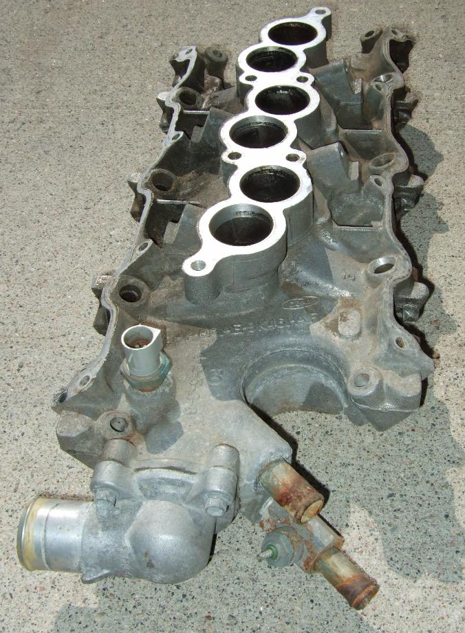 Aluminum engine intake manifold with six ports, a sensor, and a thermostat housing, resting on a concrete surface