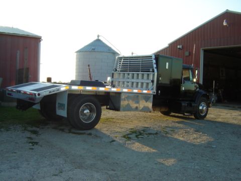 1992 Ford Bronco - Flatbed tow truck with sleeper cab parked on gravel near farm buildings.