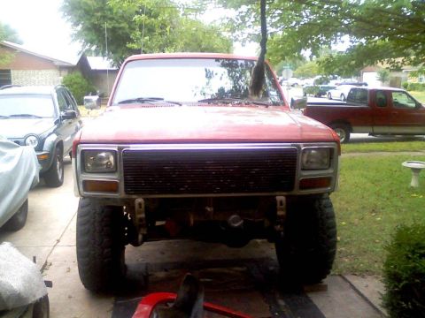 1986 Ford Bronco - This red pickup truck is parked in a driveway with other vehicles nearby. The front end is visible, showing its grille and headlights.