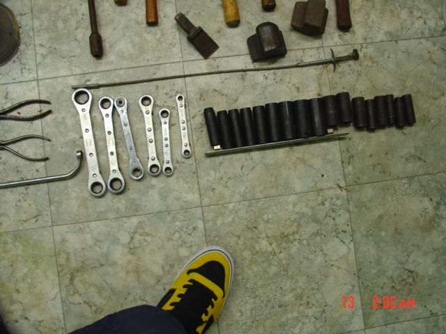 Tools laid out on a floor, including wrenches, sockets, pliers, and hammers. A person's foot in a yellow and black sneaker is visible