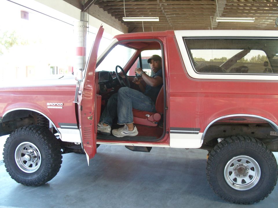 Red Ford Bronco with white trim, driver's door open, man in driver's seat. Visible text 'BRONCO' on fender