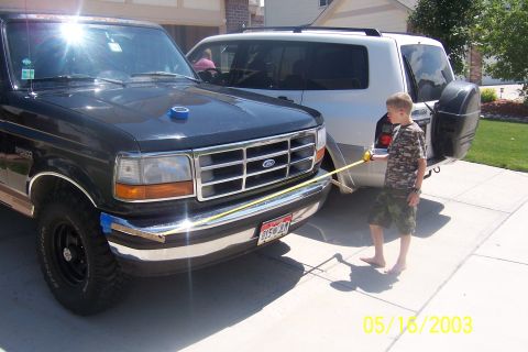 1996 Ford Bronco - A boy measures the front of a black Ford Bronco parked in a driveway next to a white SUV.